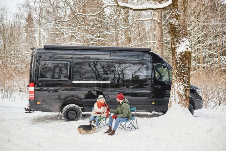 Téléchargez les photos : Portrait grand angle de jeune couple voyageant en van à l'extérieur dans la forêt d'hiver avec chien mignon, espace de copie - en image libre de droit
