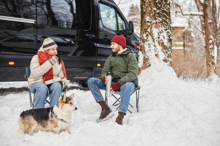 Foto de Retrato de larga duración de feliz pareja joven viajando en furgoneta en el bosque de invierno y sentado al aire libre con lindo perro corgi, espacio de copia - Imagen libre de derechos