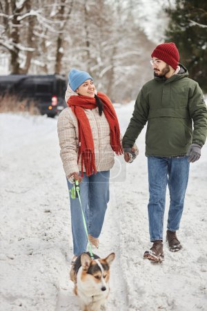 Téléchargez les photos : Portrait vertical pleine longueur de heureux jeune couple marchant mignon chien corgi dans la forêt d'hiver - en image libre de droit