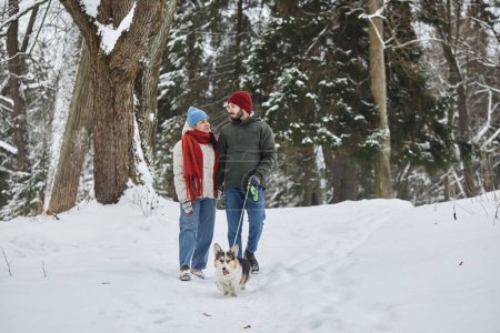 Téléchargez les photos : Portrait complet de jeune couple promenant le chien dans la forêt d'hiver et se regardant avec affection, espace de copie - en image libre de droit
