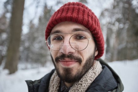 Foto de Retrato de cerca del joven barbudo con gafas mirando a la cámara al aire libre en el bosque de invierno - Imagen libre de derechos