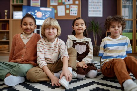 Téléchargez les photos : Vue de face d'un groupe multiethnique de petits enfants assis sur le sol en classe préscolaire et regardant la caméra - en image libre de droit