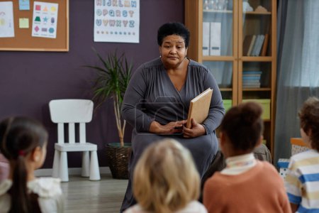 Foto de Retrato de una mujer negra amable como maestra leyendo libro a grupo de niños pequeños en preescolar, espacio para copiar - Imagen libre de derechos
