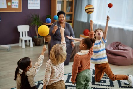 Photo for Diverse group of little kids playing with planet models while enjoying class in preschool with teacher watching - Royalty Free Image