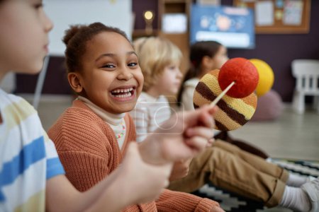 Foto de Retrato de la niña negra sosteniendo modelo planeta y riendo felizmente en la clase preescolar, espacio de copia - Imagen libre de derechos