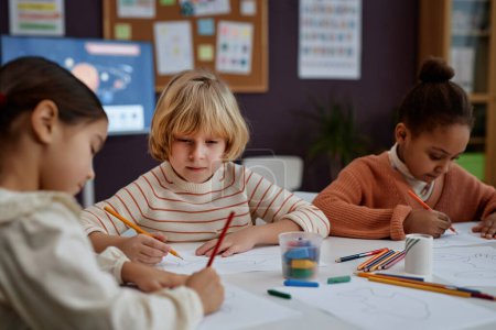 Photo for Portrait of blonde boy doing arts and crafts sitting at table with diverse group of children in preschool class, copy space - Royalty Free Image