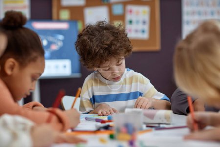 Photo for Portrait of cute boy coloring pictures at table with group of children in arts and crafts class in preschool, copy space - Royalty Free Image