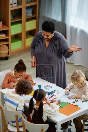 Photo for Vertical portrait of Black mature woman as female teacher helping little children in art and craft class at preschool - Royalty Free Image