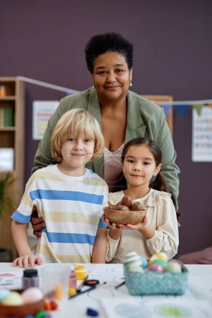 Téléchargez les photos : Portrait vertical de genre Femme noire comme enseignante posant avec de mignons petits enfants à l'école maternelle tous regardant la caméra - en image libre de droit