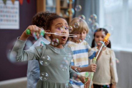 Foto de Grupo de niños pequeños y alegres jugando con burbujas de jabón en preescolar y riendo felizmente - Imagen libre de derechos