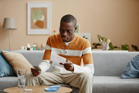 Foto de Retrato del hombre afroamericano adulto leyendo instrucciones para las píldoras recetadas mientras está sentado en el sofá en casa, espacio para copiar - Imagen libre de derechos