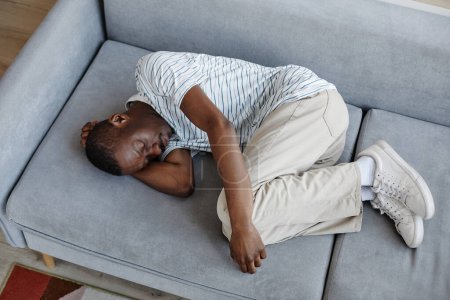 Foto de Retrato de vista superior del hombre negro acostado en el sofá en casa acurrucado en posición fetal y sufriendo de problemas de salud mental - Imagen libre de derechos
