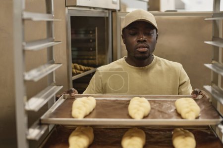Front view portrait of young Black man as worker in artisan bakery holding tray with fresh breads, copy space