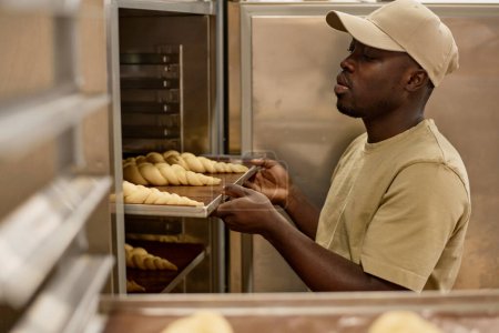 Téléchargez les photos : Portrait latéral de jeune homme noir mettant plateau dans un four industriel tout en travaillant dans la boulangerie, espace de copie - en image libre de droit