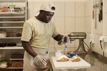Foto de Retrato de vista lateral del joven hombre negro como panadero masculino decorando cruasanes frescos con crema en panadería, espacio para copiar - Imagen libre de derechos