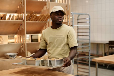 Foto de Retrato de la cintura hacia arriba del joven hombre negro sosteniendo bandeja con panes mientras trabajaba en panadería, espacio para copiar - Imagen libre de derechos
