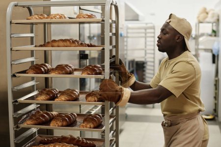 Foto de Retrato de vista lateral del joven negro moviendo estantes con panes recién horneados en la cocina de panadería, espacio para copiar - Imagen libre de derechos