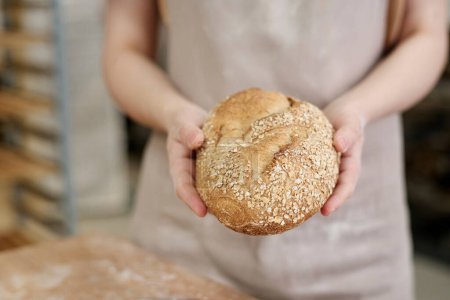 Téléchargez les photos : Gros plan d'une jeune femme tenant un pain de blé entier frais décoré de graines dans une boulangerie, espace pour copier - en image libre de droit