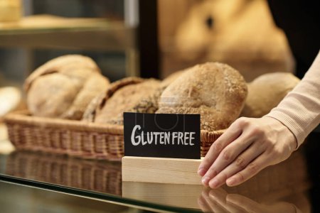 Photo for Closeup of fresh breads in artisan bakery with female hand holding gluten free sign, copy space - Royalty Free Image