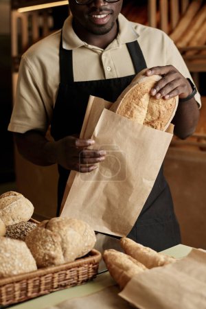 Téléchargez les photos : Gros plan vertical de jeune homme mettant du pain frais dans un sac en papier tout en servant le client dans une boulangerie artisanale - en image libre de droit