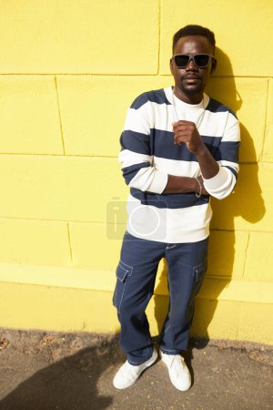 Téléchargez les photos : Vibrant portrait pleine longueur de jeune homme noir regardant la caméra posant contre le mur jaune à l'extérieur - en image libre de droit