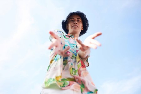 Portrait minime de jeune homme asiatique atteignant la caméra et souriant tout en portant une tenue à la mode contre un ciel bleu clair, espace de copie