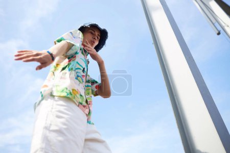 Action-Porträt eines jungen asiatischen Mannes, der vor strahlend blauem Himmel tanzt und trendiges Outfit trägt, Kopierraum