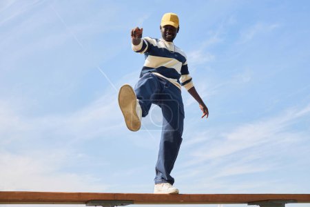 Téléchargez les photos : Prise de vue minimaliste de jeune homme noir portant une tenue tendance debout sur le toit contre le ciel bleu et donnant le coup d'envoi à la caméra. - en image libre de droit