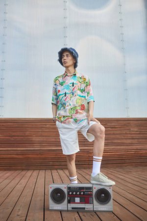 Ganztägige Modeaufnahme eines jungen asiatischen Mannes, der mit Boombox in urbaner Stadtlage posiert und ein farbenfrohes trendiges Outfit trägt
