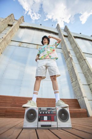 Niedrigwinkel-Modeaufnahme eines jungen asiatischen Mannes, der auf einer Boombox in einer urbanen Stadt gegen den Himmel steht