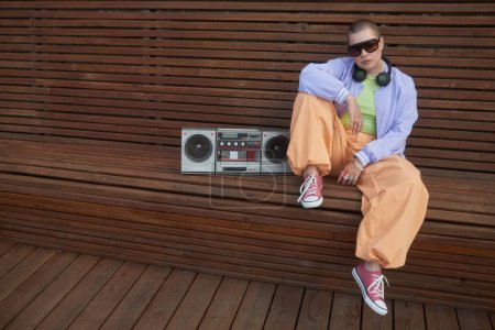 Photo for Fashion shot of bald tough girl posing with boombox sitting on bench in urban city setting and wearing trendy outfit - Royalty Free Image