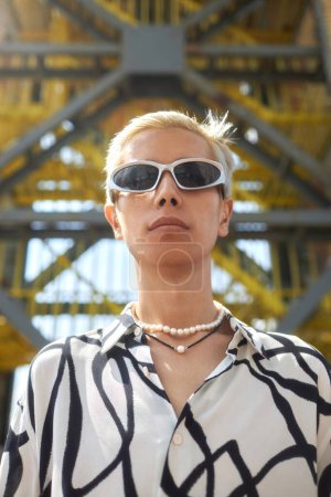 Vertikales Modeporträt eines asiatischen jungen Mannes mit Sonnenbrille und modischem Outfit, der vor einer urbanen Metallstruktur in die Kamera blickt
