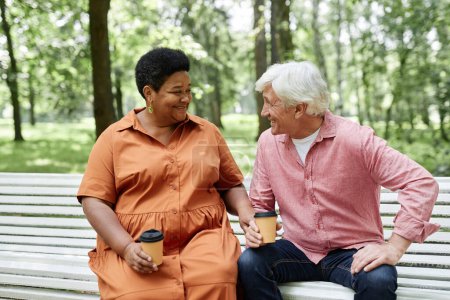 Foto de Retrato de pareja mayor multiétnica disfrutando de una cita al aire libre y sentada en un banco de madera en el parque, espacio para copiar - Imagen libre de derechos