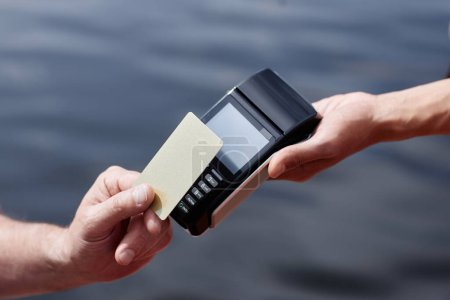 Foto de Primer plano de la persona irreconocible que paga a través de NFC con tarjeta de crédito al aire libre con agua en el fondo, espacio de copia - Imagen libre de derechos