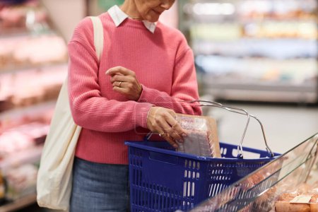 Close up of adult woman putting products in shopping basket at supermarket, copy space