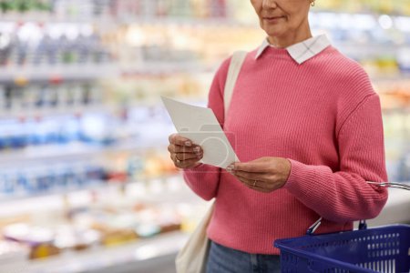 Close up of adult woman reading shopping list in supermarket and holding basket, copy space