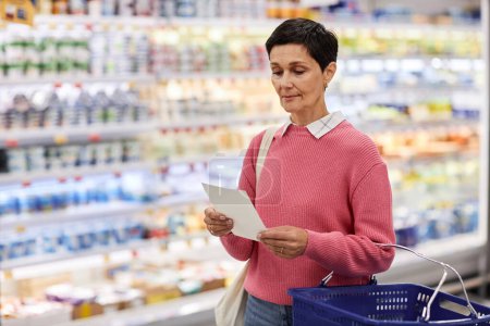 Portrait taille haute de femme adulte lisant la liste d'achats au supermarché et tenant le panier, espace de copie