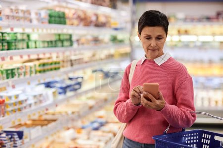 Foto de Retrato de la cintura hacia arriba de la mujer adulta usando el teléfono inteligente en el supermercado y la cesta de celebración, espacio de copia - Imagen libre de derechos