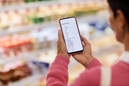 Foto de Primer plano de la mujer que sostiene el teléfono inteligente y la lectura de la lista de compras en el supermercado, espacio de copia - Imagen libre de derechos