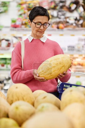 Vertical waist up portrait of woman holding melon while choosing fruits in supermarket