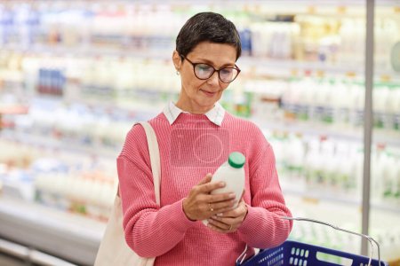 Foto de Retrato de la cintura hacia arriba de la mujer madura sosteniendo la botella de leche en el supermercado y mirando la fecha de caducidad, espacio de copia - Imagen libre de derechos