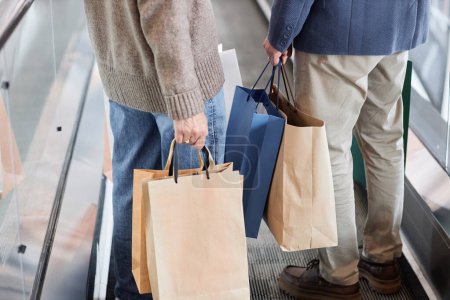 Foto de Recortado disparo de pareja sosteniendo bolsas de compras de papel mientras está de pie en la escalera mecánica en el centro comercial, espacio de copia - Imagen libre de derechos