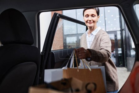 Photo for Portrait of smiling adult woman loading paper bags in car outside shopping mall, copy space - Royalty Free Image