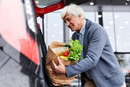 Foto de Retrato de vista lateral de hombre mayor sonriente poniendo bolsas de comestibles en el maletero del coche al aire libre, espacio para copiar - Imagen libre de derechos