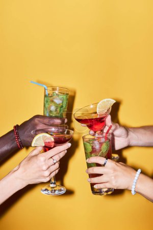 Foto de Primer plano vertical de grupo multiétnico de amigos sosteniendo vasos de cóctel sobre fondo amarillo y tostadas, espacio para copiar - Imagen libre de derechos