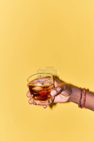 Photo for Vertical closeup of male hand holding whiskey glass on vibrant yellow background, copy space - Royalty Free Image