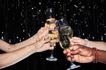 Photo for Closeup of hands toasting with champagne glasses at party against glittering background with confetti - Royalty Free Image
