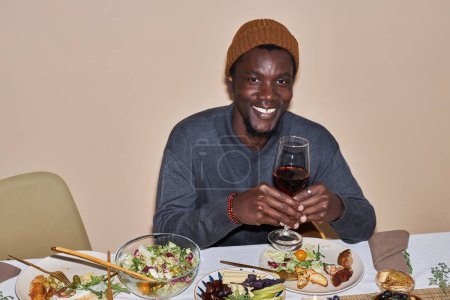 Photo for Portrait of young Black man smiling at camera and holding wine glass at dinner table for Thanksgiving, copy space - Royalty Free Image