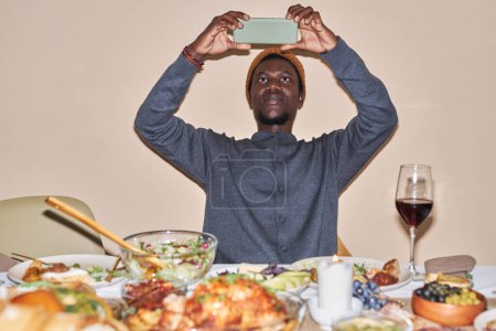Photo for Front view portrait of young African American man taking photo of Thanksgiving dinner table shot with flash, copy space - Royalty Free Image