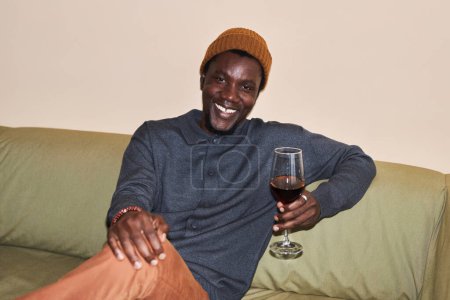 Foto de Retrato de joven negro confiado con copa de vino tinto sentado en el sofá sonriendo a la cámara, filmado con flash - Imagen libre de derechos